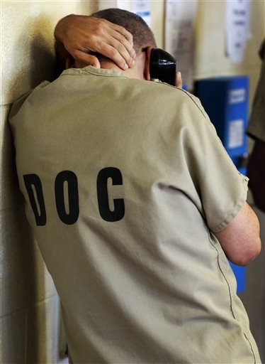 An inmate in jail is photographed facing away from the camera leaning against a concrete wall with his hand around his head while talking on the phone. 