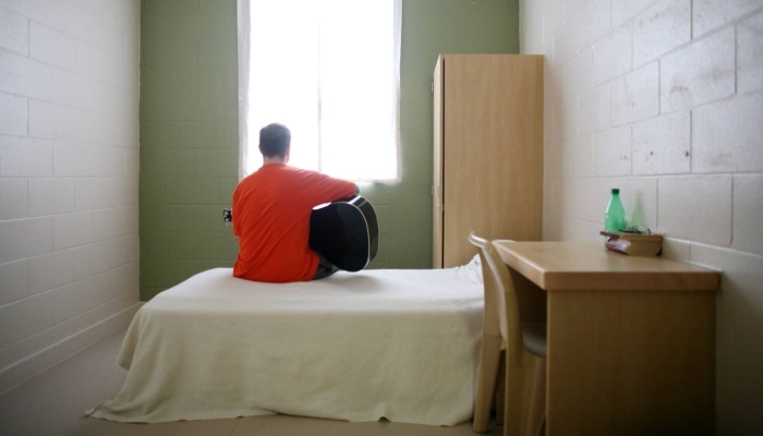 Inmate in his cell sitting on his plain white bed looking out his window while playing guitar. 